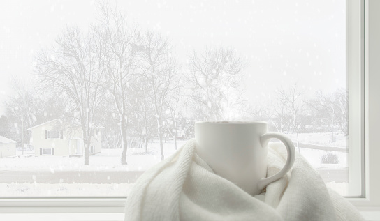 Hot tea mug covered by white comfort soft wool scarf in front of frosted glass window in warm and cozy living room with lonely but beautiful landscape view snow falling outside.